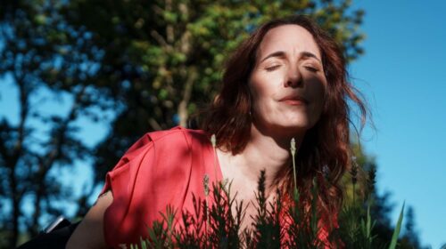 Imagem de uma mulher em uma área externa e arborizada. Ela é branca, em os cabelos ruivos e veste uma blusa cor de goiaba. Ela está cheirando flores de lavanda.