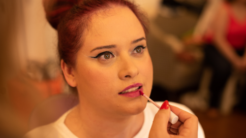 Imagem de uma mulher branca de cabelos vermelhos. Ela está maquiada e com a boca entreaberta e uma mão externa segura o aplicador de batom perto de sua boca.