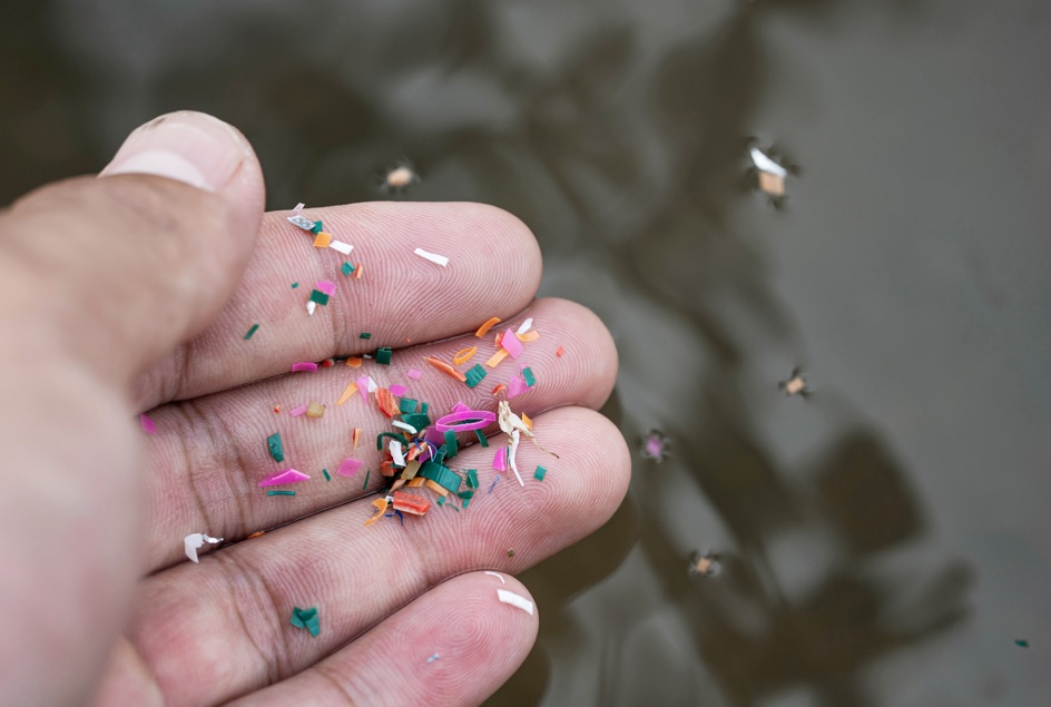 Imagem da mão de uma pessoa segurando pedaços de microplásticos. Ao fundo da mão, água.