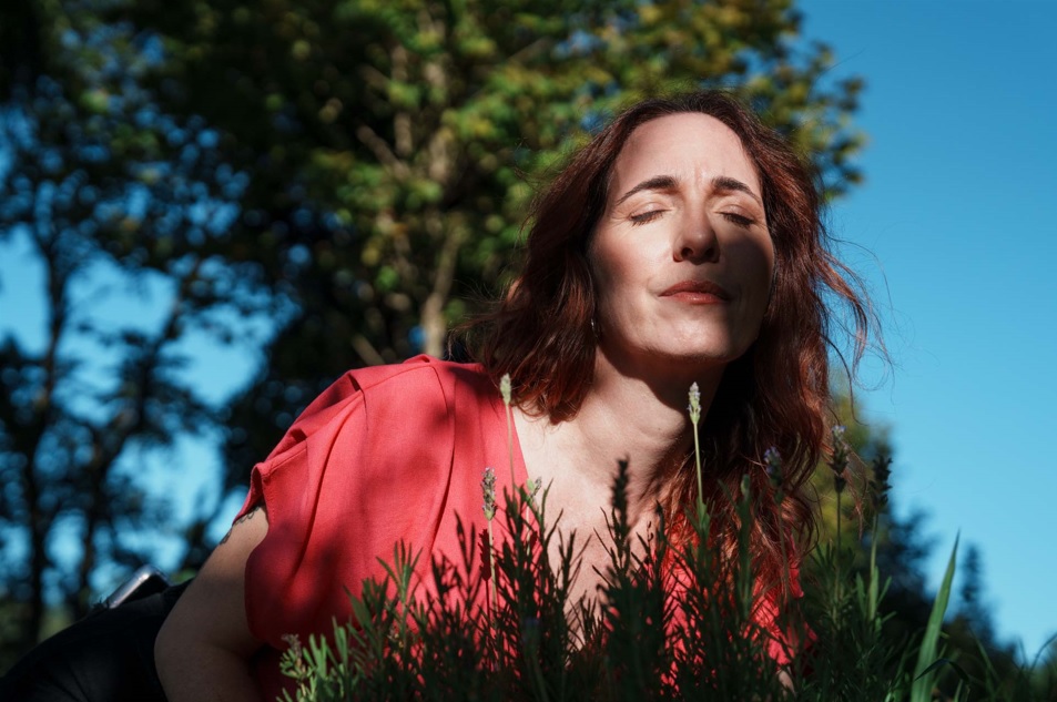 Imagem de uma mulher em uma área externa e arborizada. Ela é branca, em os cabelos ruivos e veste uma blusa cor de goiaba. Ela está cheirando flores de lavanda.