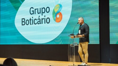 Imagem de um homem em frente ao púlpito, no palco do Congresso de Cosmetologia IFSCC. Ao fundo, no telão, logo do Grupo Boticário.