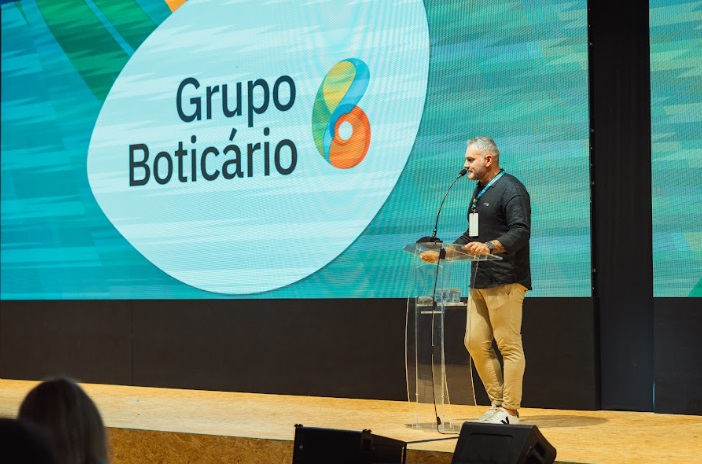 Imagem de um homem em frente ao púlpito, no palco do Congresso de Cosmetologia IFSCC. Ao fundo, no telão, logo do Grupo Boticário.