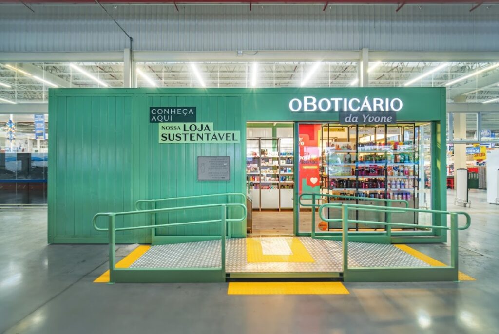 Imagem da fachada de uma loja container de O Boticário. O container é verde e, do lado esquerdo da fachada está o texto "Conheça aqui nossa loja sustentável". À direita, acima da porta de entrada, está o letreiro "O Boticário da Yvone". A ação faz parte da homenagem da empresa às mulheres empreendedoras.