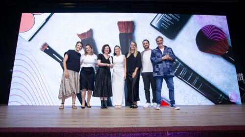 Imagem de sete pessoas em cima de um palco, são cinco mulheres e dois homens, sendo que duas mulheres seguram um troféu. Ao fundo, no telão, está projetada uma imagem dos pincéis acessíveis de Make B., de O Boticário.