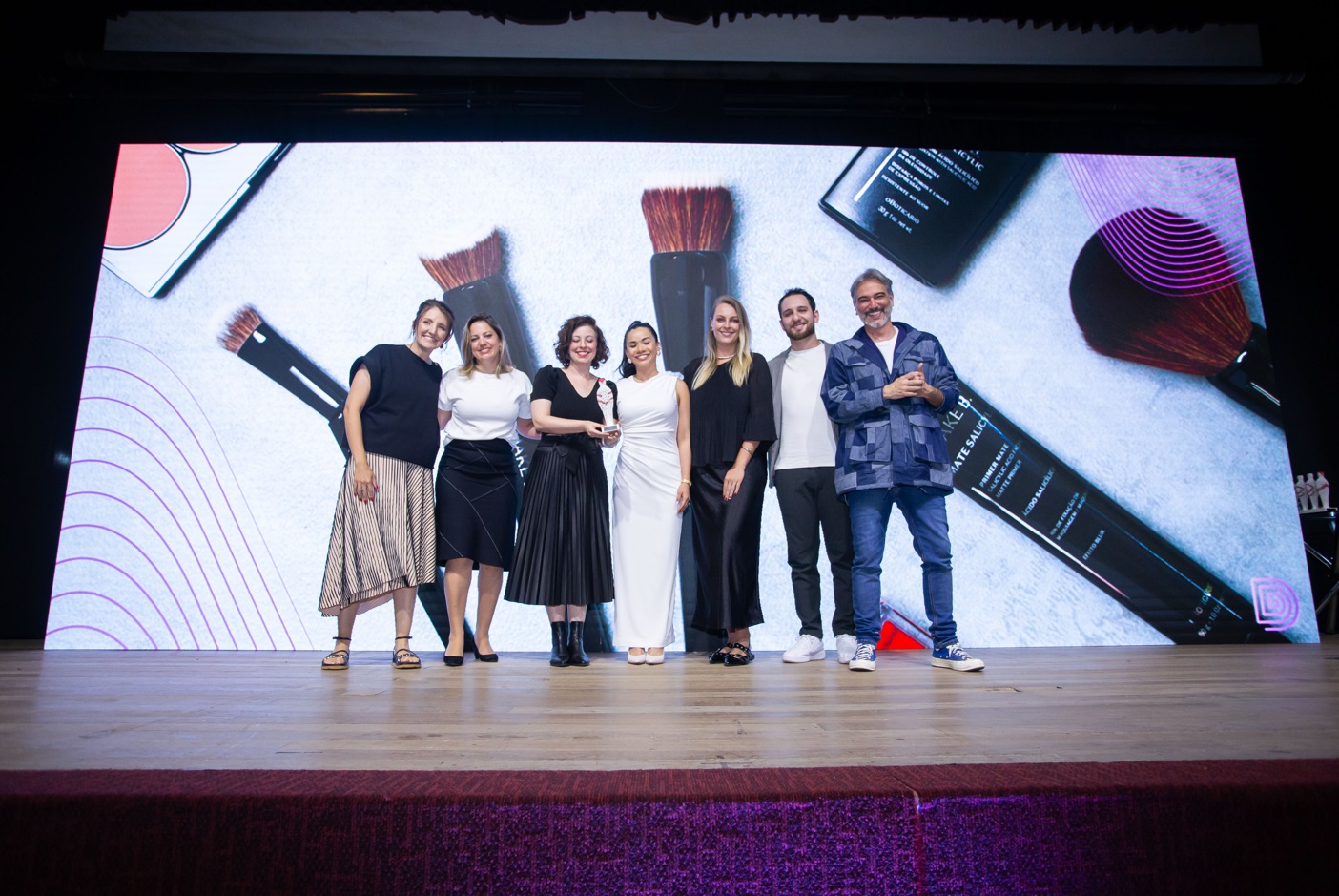 Imagem de sete pessoas em cima de um palco, são cinco mulheres e dois homens, sendo que duas mulheres seguram um troféu. Ao fundo, no telão, está projetada uma imagem dos pincéis acessíveis de Make B., de O Boticário.